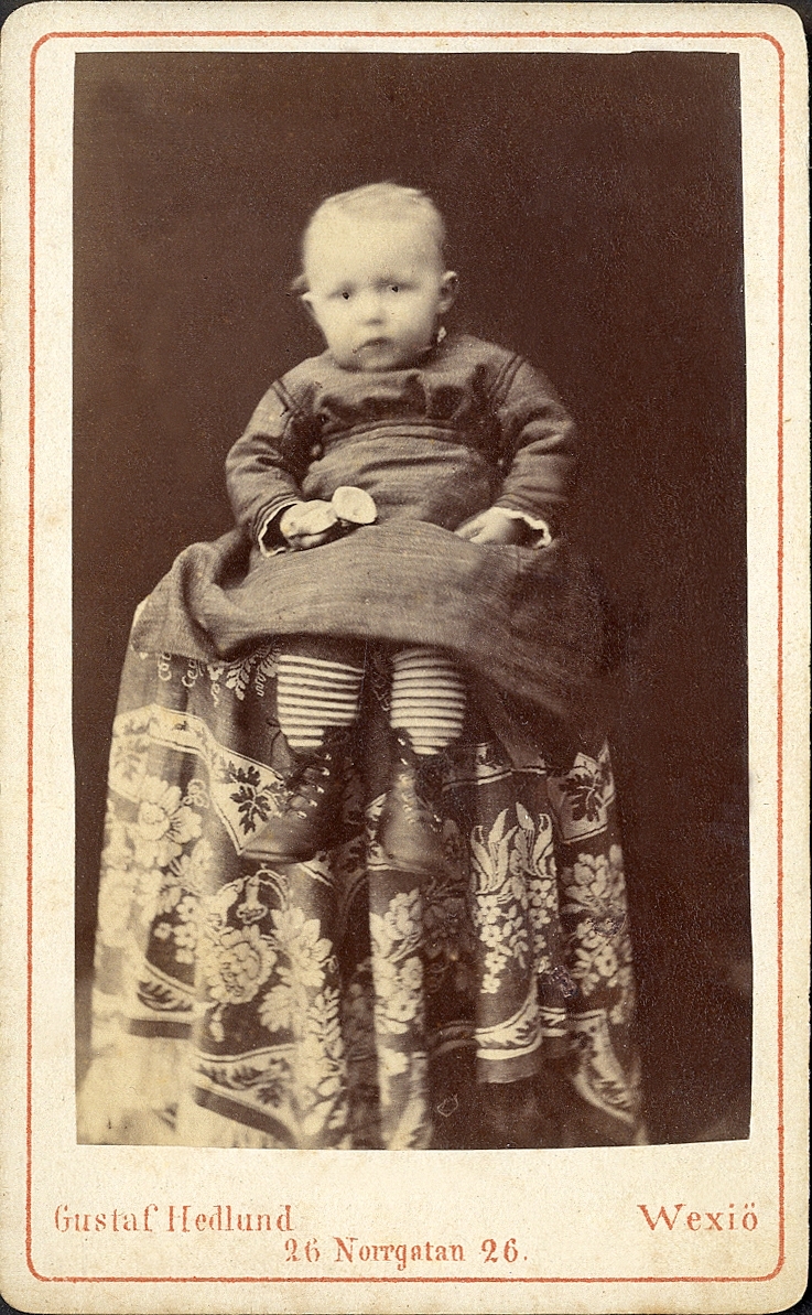 En liten pojke(?) i kolt och tvärrandiga strumpor, sitter uppflugen på ett bord med en blommig duk.
Helfigur, en face, Ateljéfoto.