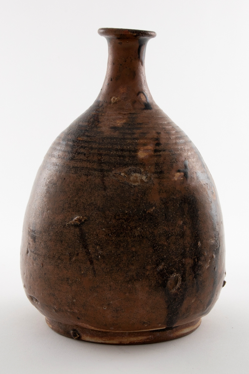 Pæreformet vinflaske med trang hals og utbrettet kant. Foten er vid med bred rand. Flasken er glasert i brun med nyanserte sjatteringer.