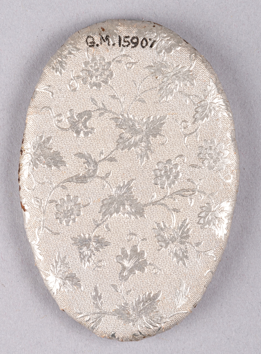 Begravningskonfekt, platt, oval konfekt, klädd med mönstrat silverpapper. Ovanpå garnerad med vit tyll och en konstgjord liljekonvalj.