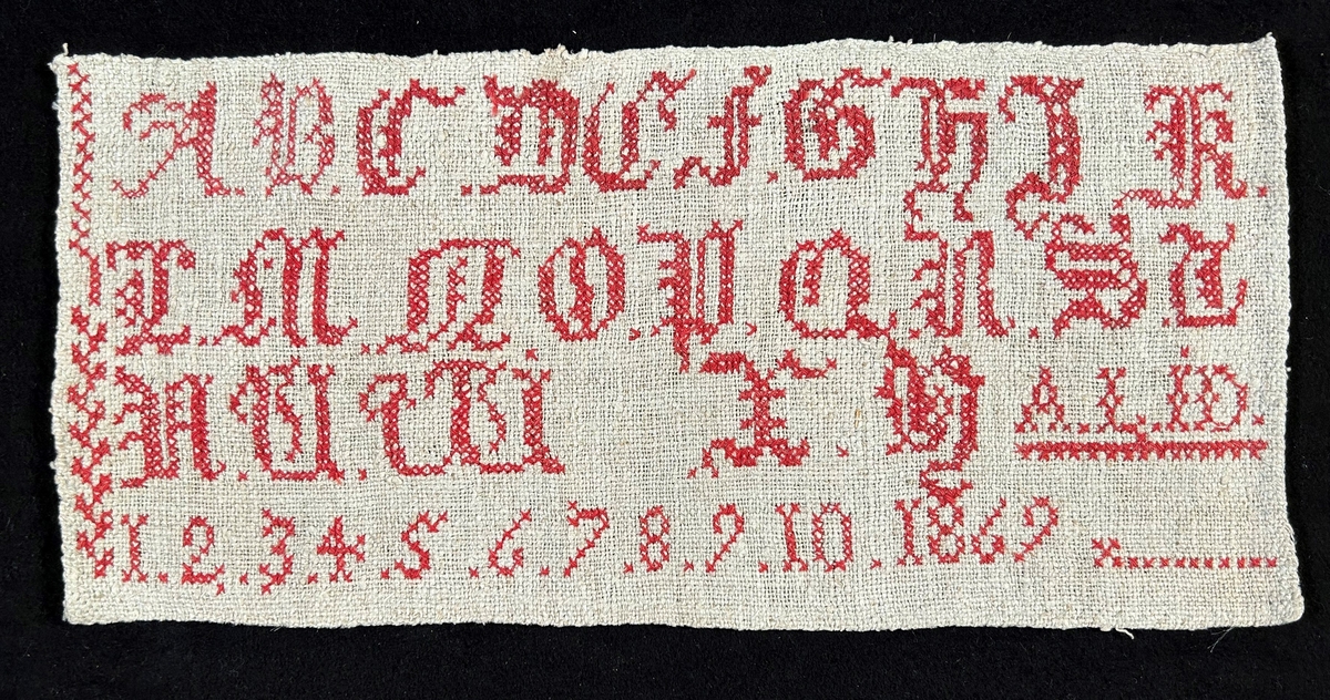 Märkduk i grått linne med korsstygnsbroderi i rött: Alfabetet A-Y, siffror 1-10 1869, samt initialerna A. L. - I. D.