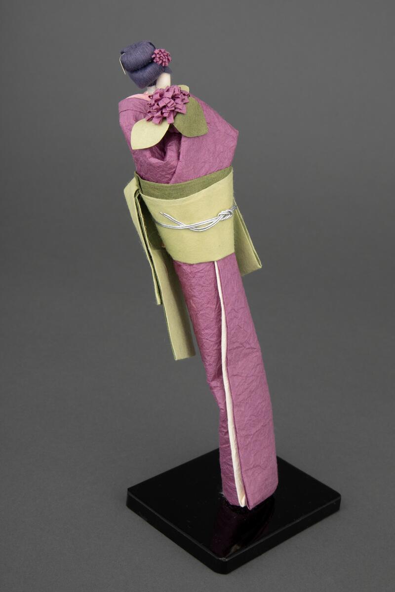 Håndlaget papirdukke i japansk bretteteknikk plassert på en svartmalt tresokkel. Alle deler er laget i papir bortsett fra "kroppen" som består av en trepinne og vatt. Dukken er iført en lilla kimono og har oppsatt hår med lilla blomst. I henden har hun en noe større, lignende blomst. Hun har rosa innerkrage og hvit innerkimono og mørk- og lysegrønn obi med sølvfarget obisnor.