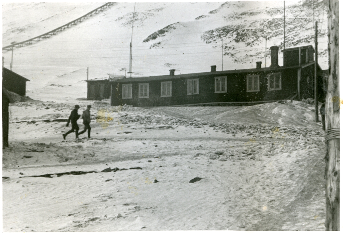 To menn på vei forbi en boligbrakke i  Longyearbyen i 1940.  Nesten alle husene der ble brent av tyske soldater i 1943.  Bilde fra albumet til Jens A. Bay, som bodde i Longyearbyen vinteren 1940/41.