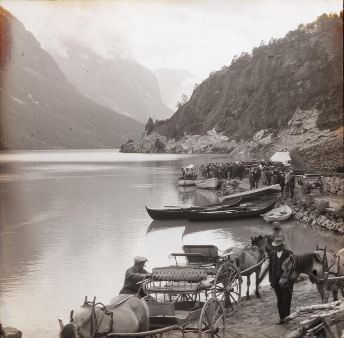 Dias. To menn står med vogner spent for fjordinger. I bakgrunnen sees en gruppe kvinner og menn på en brygge.