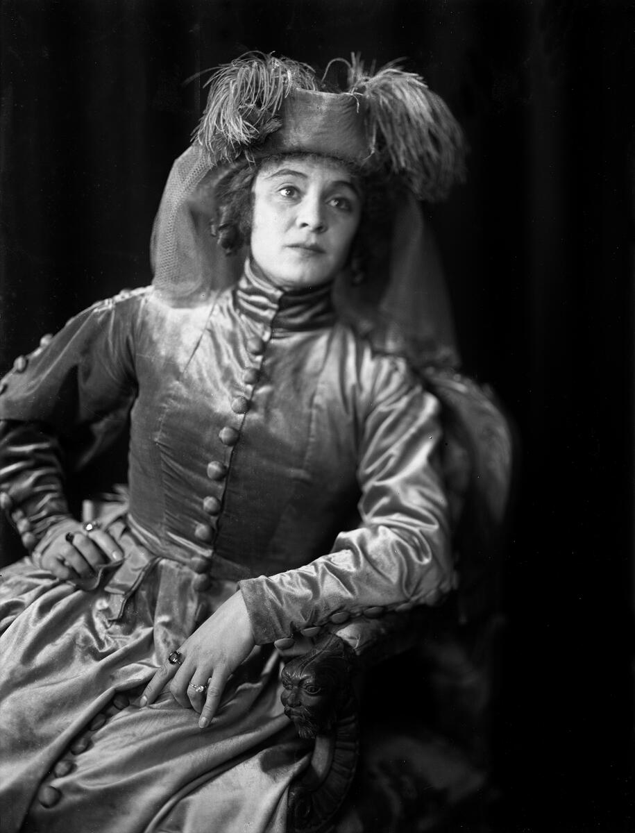 Portrett av skuespilleren og sceneinstruktøren Gerda Ring. Hun poserer sittende i en lenestol ikledd en langermet kjole med knapper og hatt med fjær og slør.
