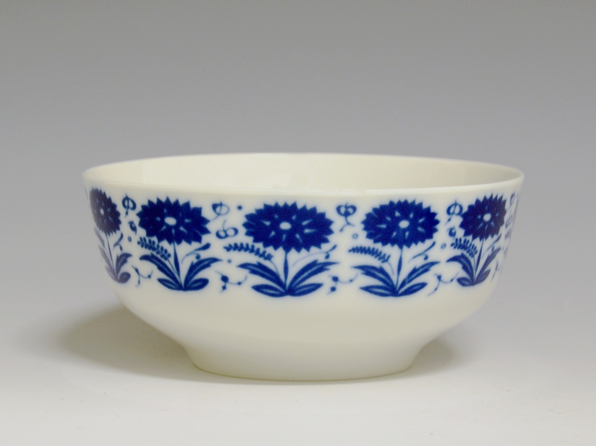 Sausebolle med fat, av porselen. Dekorert med blomsterdekor i blå iglasur trykkdekor.
Modell: Meny, 2310 av Tias Eckhoff, i produksjon fra 1955.
Dekor: Nellik av Anne-Marie Ødegaard fra 1957.
