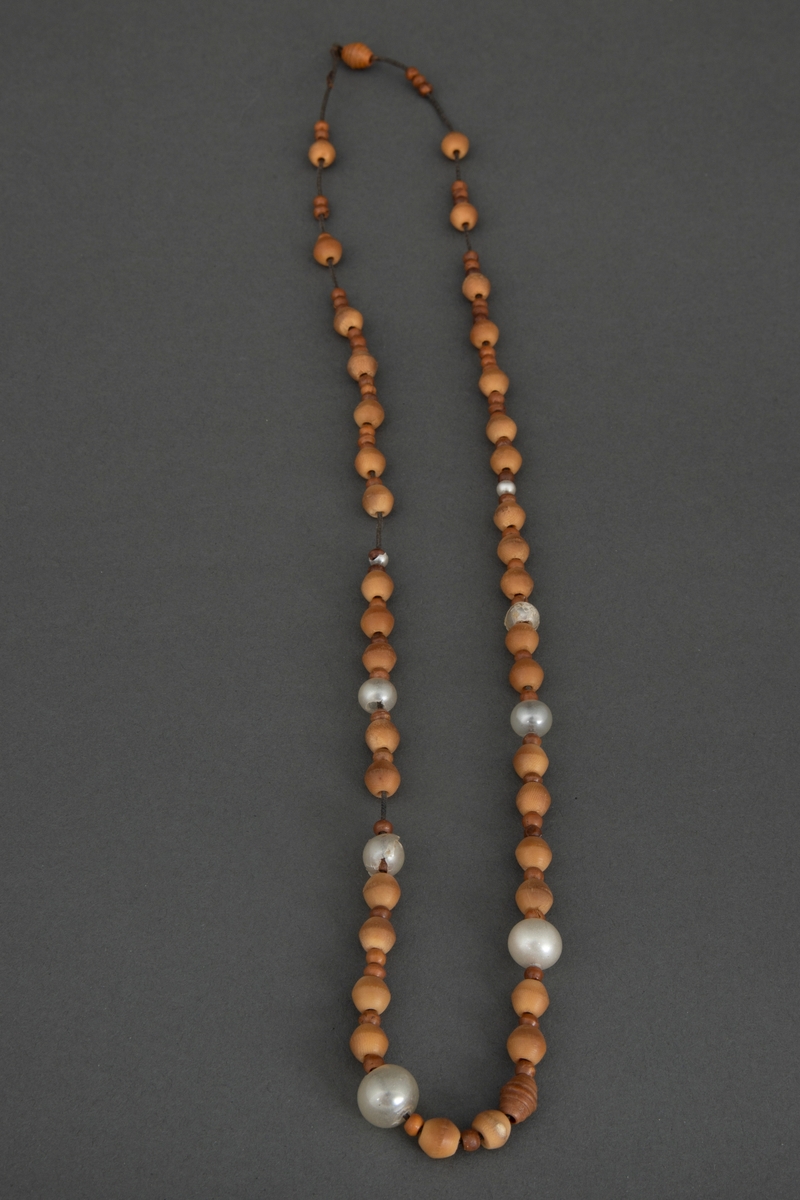 Perlehalssmykke bestående av større og mindre, dreide benperler, lysebrune og brune, og noen hvite glassperler. Perlene er tredd på en svart, noe elastisk tråd som har strukket seg sterkt.