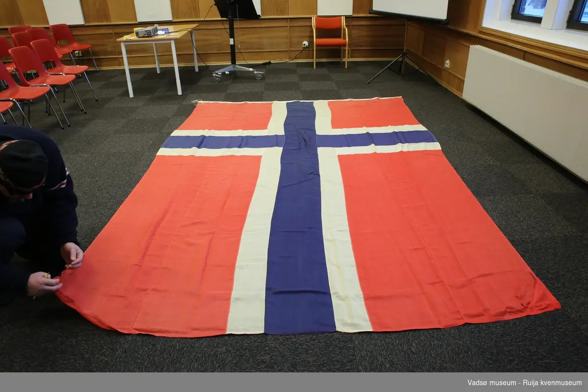 Norsk flagg i vevd materiale, antatt syntetisk. Løpegang og snor av bomullstoff. Flagget har maskinsydd søm, hvitt og rødt er brukt.