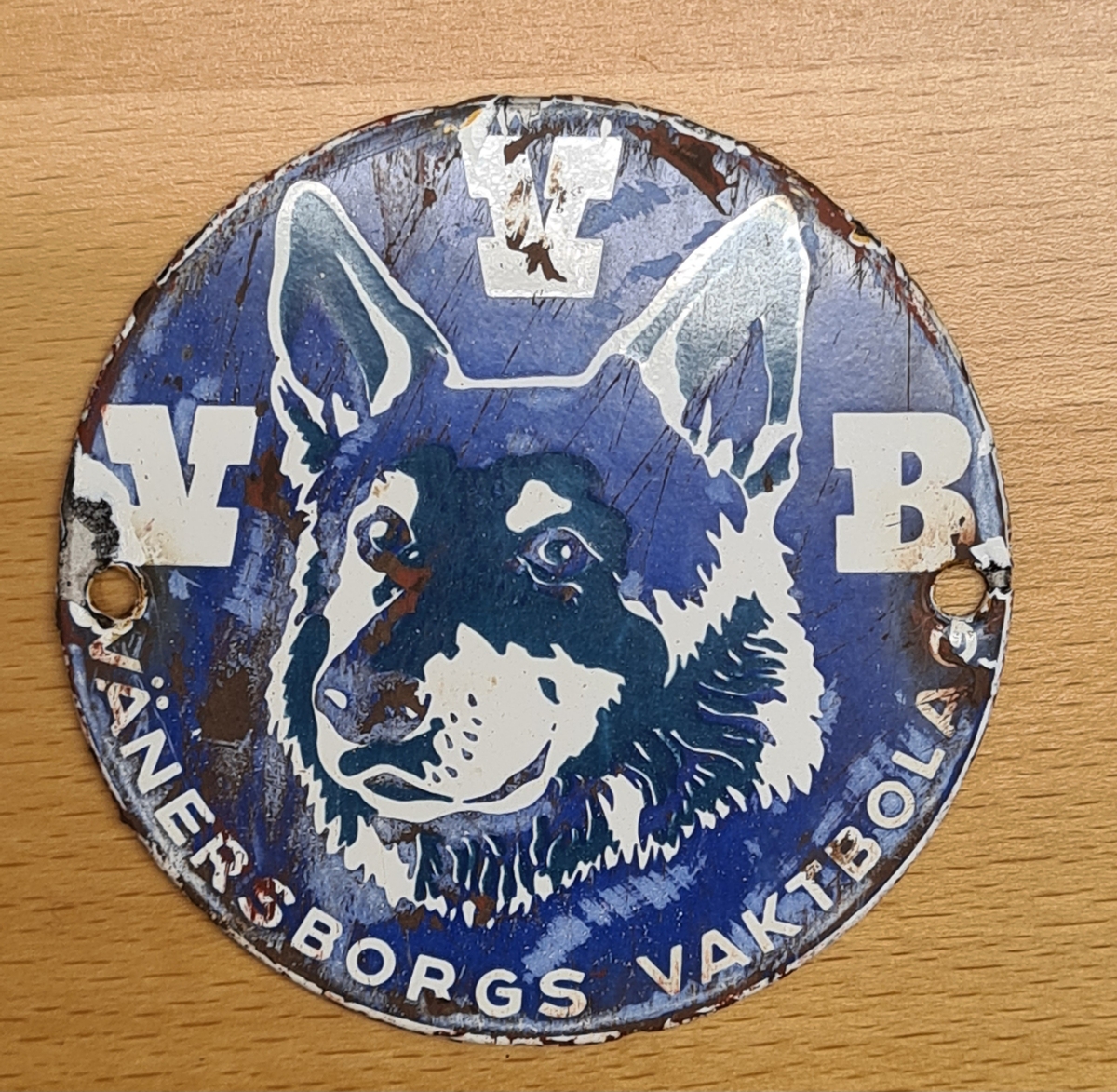 Skylt från Vänersborgs vaktbolag (VVB). 
Det är en rund skylt med två hål för montering. Skylten är emaljerad med en dekor med en hund.
Skylten var monterad på Vänersborgs museums dörr. Den framkom då dörren skrapades inför en ommålning.