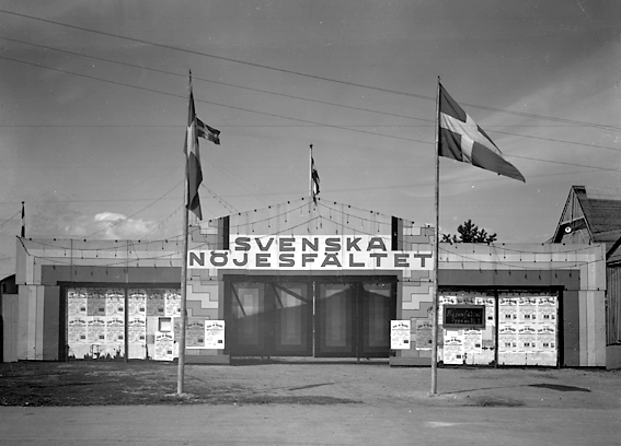 Entrén till Svenska nöjesfältet med flaggstänger som flankerar ingången. Text på skylt: Nöjesfältet öppnas Kl 7.