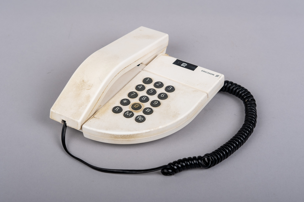 Ericsson bordtelefon.
Apparaten består av telefonrør, teelfon og spiralkabel. 
Hvit telefon med 15 svarte trykk knapper: tall (0-9), stjerne, nummertegn, funkjsonsknapper. 

Telefonen er misfarget, brune bruksmerker på telefonrør og rundt knappene, spesiell ved tallet 0.

På bunnen klistemerke med artikkel kode:
DBCN 2120700/987 R1A
ETM 9107 D. Design
