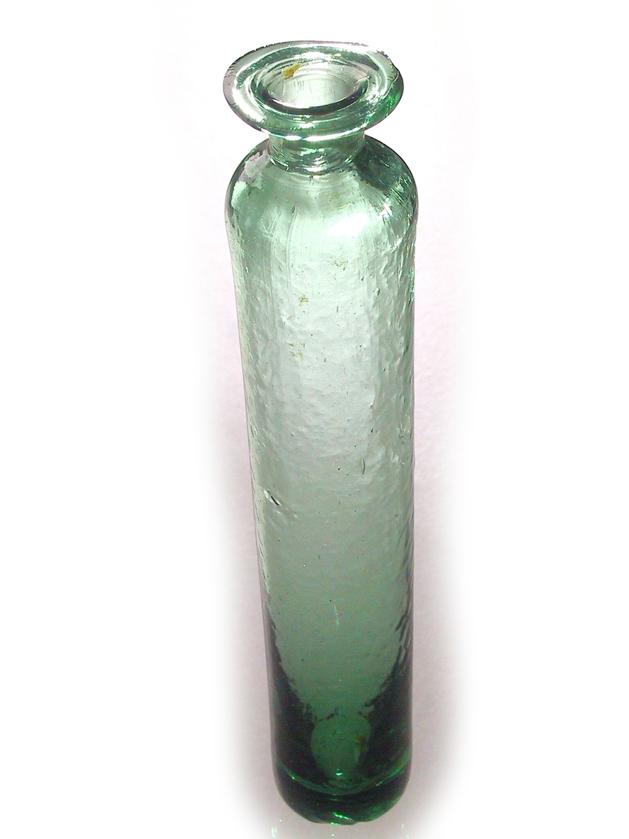 Medisinglass, Nøstetangen eller Biri. Grønt glasst. Lang smal sylinderform, kort hals og utbrettet munningsrand. Ruglet glass. Et lite skår i bunnen.