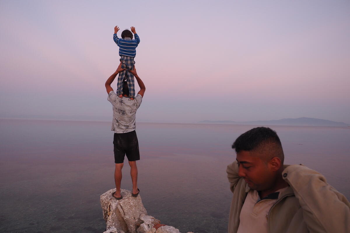 En stille kveld på stranden ved Pikpa med utsikt mot Tyrkia. En far står med sønnen på skuldrene og en annen person står i forgrunnen.