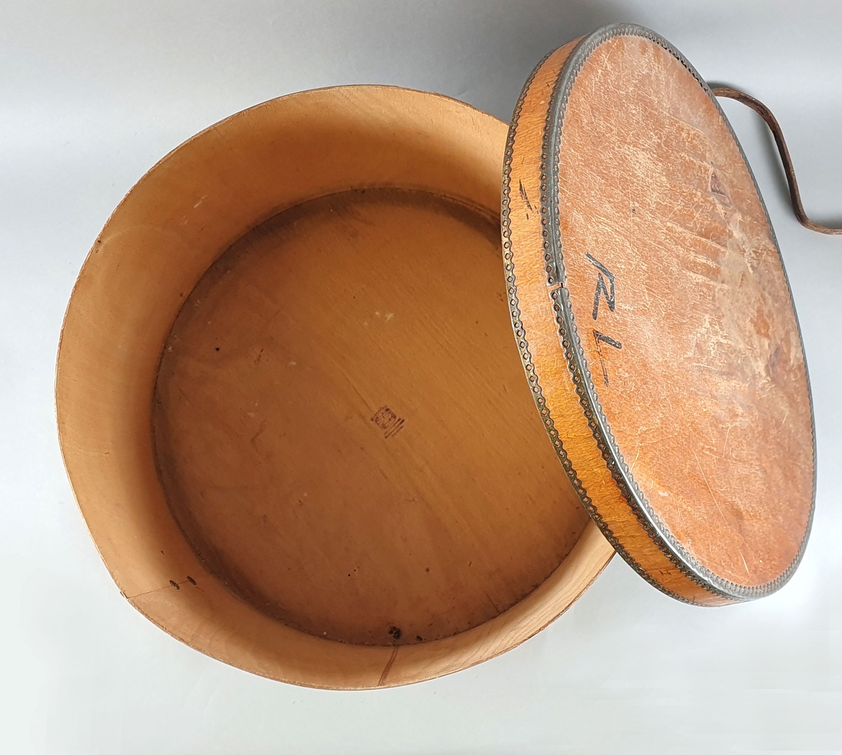 Stor, sylinderformet hatteeske av finer, forsterket med metall rundt kantene. Skinnreim med spenne over lokket, festet på hver side av esken.