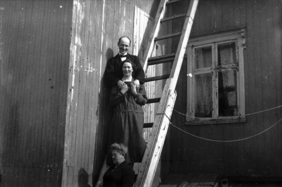 Portrett av to kvinner og en mann, fotografert i en stige ved en husvegg.