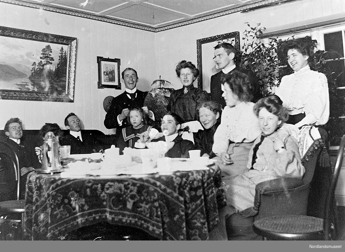 Tolv personer som sitter i en stue på Nes i Lødingen og drikker kaffe. Alle personene på bildet er finkledde. Et bord står i forgrunnen, dekket med kaffekopper, kakefat og kaffekanne.