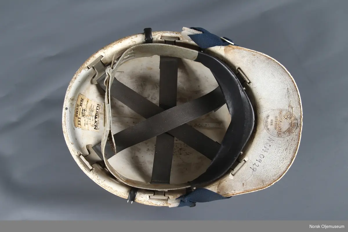 Vernehjelm med Norwegian Contractors sin logo og klistremerke med brukers navn foran. Hjelmen har opprinnelig hatt påmontert hørselsvern.