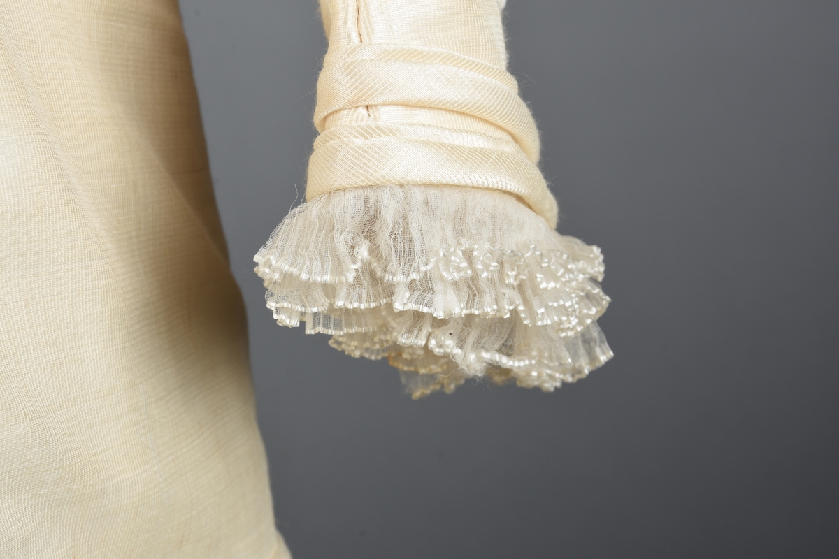 Tournure-kjole av perlehvit, gasvevd natursilke. Helfôret i livet med tynt lerret, i ermene med dunlerret, og i skjørtet med stivet batist.  
Foran er kjolen delt i en lang figurjakke over et skjørt med «paneleffekt». Jakken er lukket med tretten stofftrukne knapper som har en lav kuppelform. Knappene er også konsentrisk oppdelt med topp og kant i kjolestoffet og beige midtrand. 
Smale ermer med tre knapper ved håndleddene. Ermer og hals er kantet med tredobbelt tyllrysj og silke-roulorer. Jakken er kantet nede med tungeknipling og roulorer. 
Skjørtet er panelt med vannrette rynker under syv loddrette roulorer. På hver side av panelet er det en utbrettet flik kantet med tungeknipling og roulerer. Skjørtekanten har en 12 cm bred, tett foldet og tverrsydd rysj av kjolestoffet. Stikklomme ved høyre side av panelet. Oppunder jakken er panelet erstattet av batistfôr. Innvendige opphengsstropper i ermegapene. 
Ryggen er helskåret figur. Midtsømmen åpner seg til motfold over setet. Kantet nede med tyllrysj. Innvendig er det fire rekker á fire bendelbånd til å hefte skjørtet sammen og opp til tournure (tre bånd mangler). Noen av båndene er knyttet sammen på tvers og trekker skjørtet sammen.
Ufullendt forandring (inntaking) øverst på skjørtets forside. Det er uvisst om sammenbindingene av tournure-båndene er korrekte/originale eller senere forsøk.