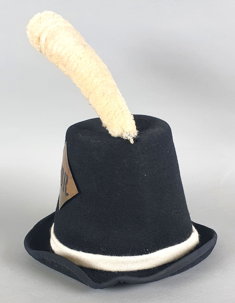 Svart hatt av filt med skinnreim på innsiden og med hvit dusk av ull på toppen og et skilt av metall og et hvit bånd av ull rundt pullen. Hatten er en kopi av en offisershatt.