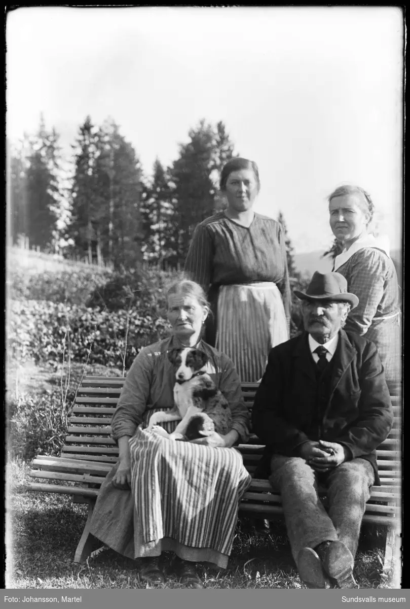 Makarna Andrietta och Johan Persson sitter på en bänk i trädgården, bakom till höger deras dotter Sigrid och till vänster om henne Hildur Grafström. Andrietta har en hund i famnen.