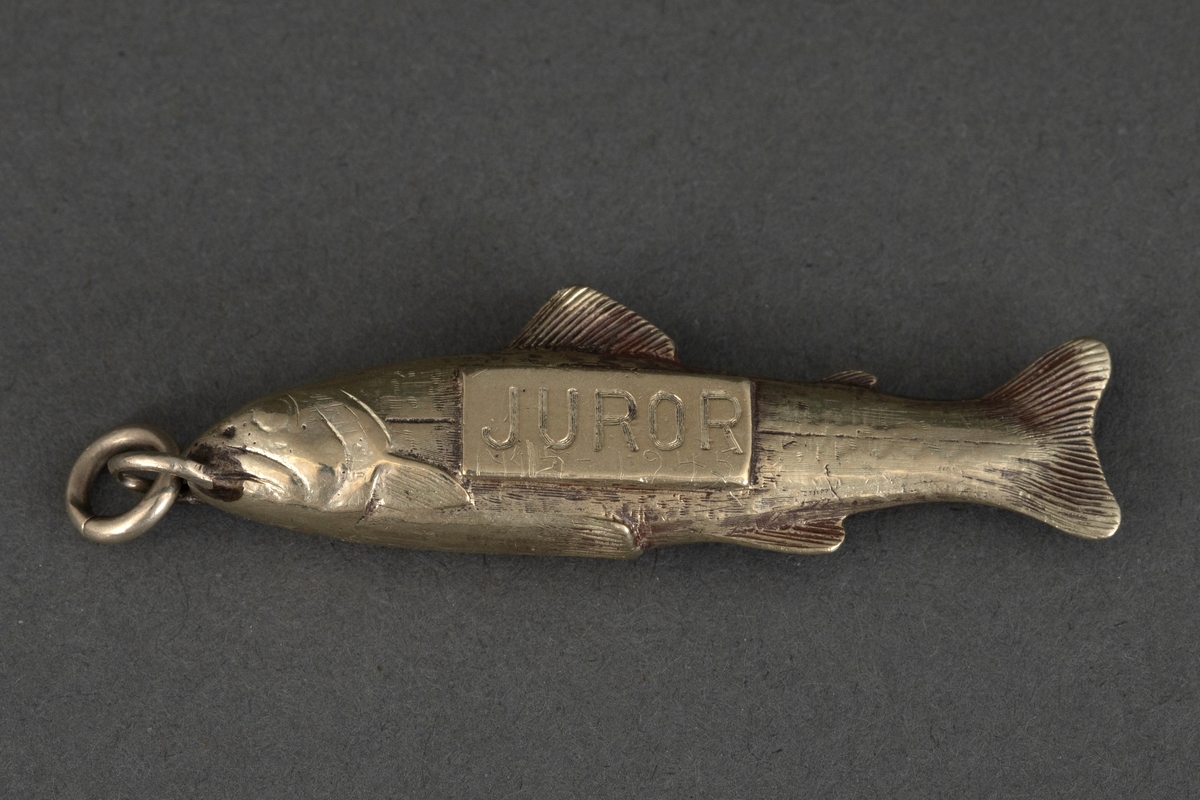 Fiskeformet merke for jurymedlem ved The International Fisheries Exhibition 1883. Hvit metall, antagelig forsølvet. Ring for feste til kjede eller lignende.
