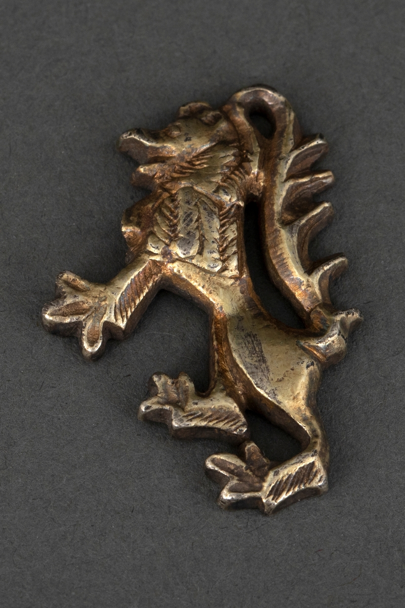 Del av spenne i forgylt sølv, i form av en løvefigur med sterkt svunget hale, stående på bakbenene. Har ifølge katalogen vært en av to flankerende figurer omkring en plate.