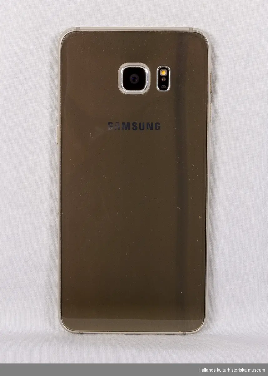 Samsung Galaxy S6 Edge+ (Tillverkare: Samsung, modell: Galaxy S6 Edge+). Grå metallic. Båda sidorna av kameran är täckta med ett skyddande lager armerat plast. Sidor av metall. 

På telefonens framsida en digital skärm (välvd vid kanterna - därmed namnet Edge) med sensor för tryck, en tryckknapp, högtalare, kameraoptik, samt en märkning: Samsung. 

På telefonens baksida kameraoptik samt två märkningar: "Samsung" och "CE0168. SM-G928F SAMSUNG. YATELEY, GU46 6GG. UK. MADE BY SAMSUNG. IMEI: 353121/07/108245/4 S/N:R58G80VTG4N"

På telefonens sidor finns diverse tryckknappar och uttag. På telefonens ovansida en lucka för telefonkort (SIM).