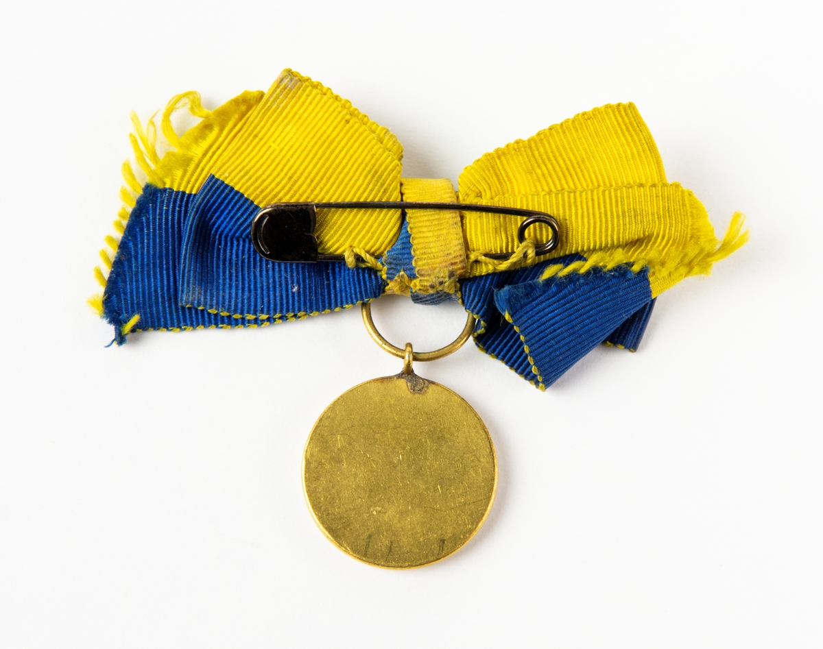 Medalj, Röda Korset med blågul rosett. Medaljen fästes på baksidan med en säkerhetsnål.