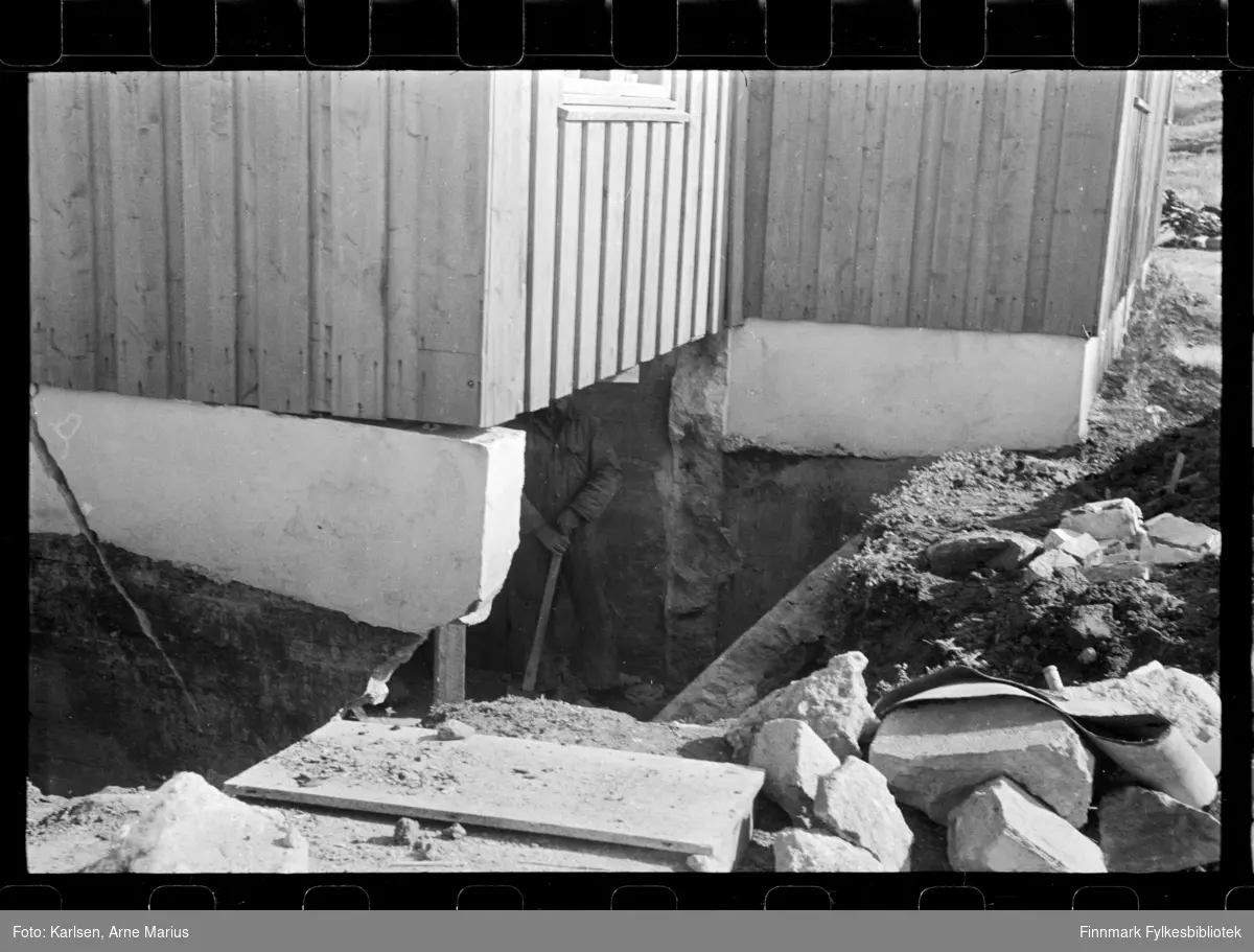 Skadet grunnmur på hus i Kirkenes 

Man kan se en mann med spade under huset 

Foto antagelig tatt på slutten av 1940-tallet, tidlig 1950-tallet 