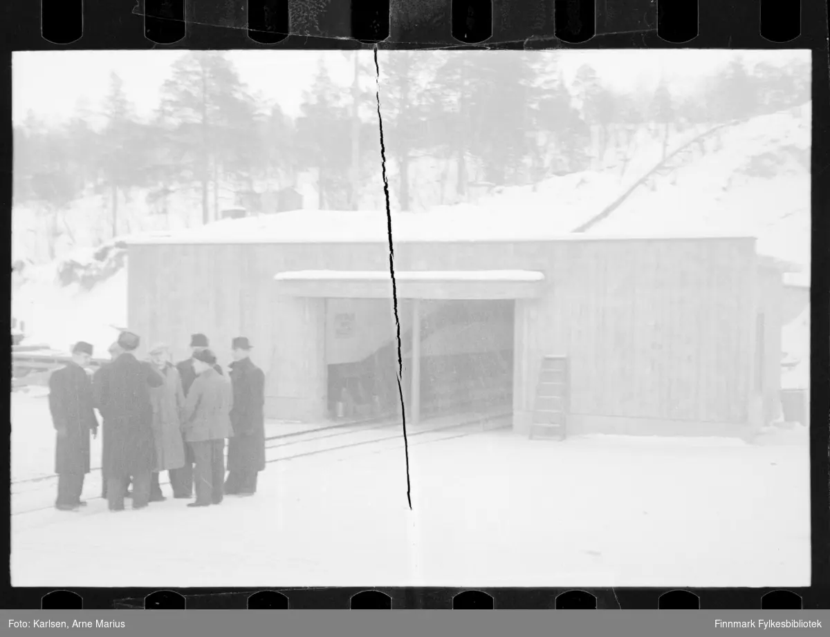 Ukjente menn står foran gruveinngang i Nikel/Kolosjoki gruve. Fotografen reiste sammen med flere andre personer til det som tidligere var Finland til Salmijärvi og Nikel før krigen i 1938. Antagelig er fotografiet tatt under denne turen (se historikk for referanse). I notatene tilknyttet negativ samlingen nevnes Bjarne Rist og Bjarne Hatle som deltakere på turen. Hvem de er av mennene er for øvrig uklart. 

Nikel er i dag russisk område 