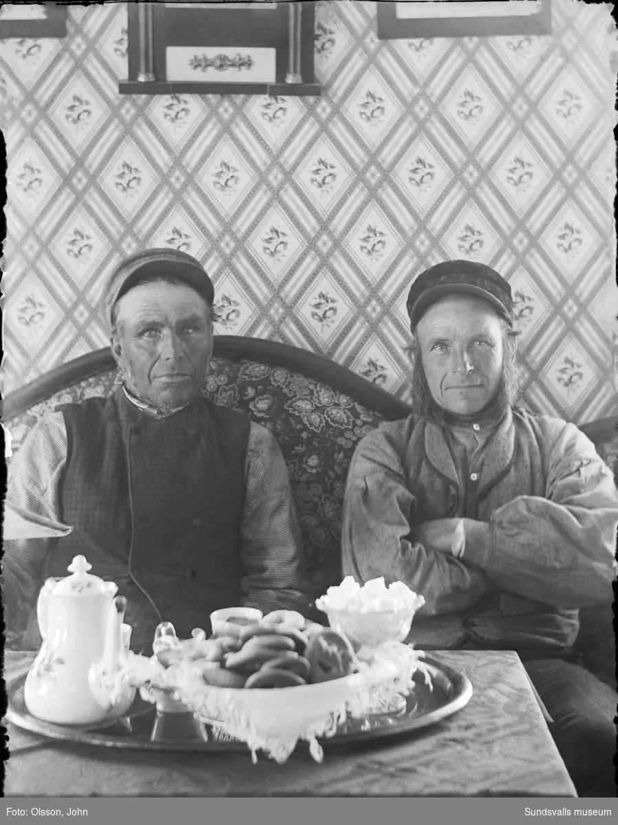 Porträttbild inomhus med fotografens bror Olof Olsson och grannen Petter, "där väst vid sjön i Ingeborgjorden" (Sunne, Värmland). På bordet framför står kaffebrickan dukad.