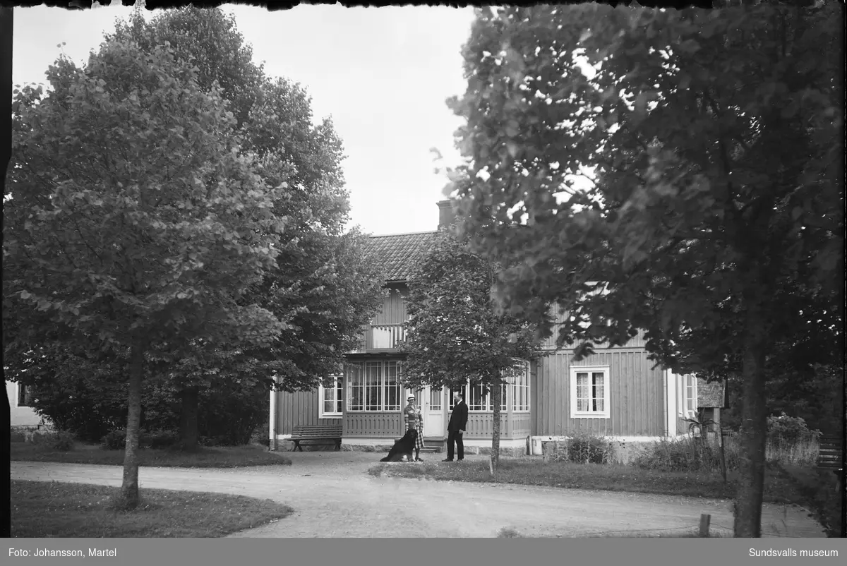 Två bilder på en bostadsbyggnad med trädgård. Framför glasverandan står en kvinna med en stor hund samt en man som möjligen är fotografen själv, Martel Johansson. Den ljusa byggnaden som skymtar i vänsterkanten är samma som SuM-foto034836, Villa Göta. Okänt var i Sverige bilden är tagen.