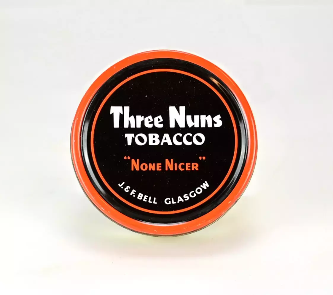 Plåtask. Three Nuns Tobacco "Non Nicer". J. & F. Bell Glasgow. 50 grammes net weight. Orange/svart/vitt lock. Etikett: "Import Riktpris 5:70 kr". Präglat "024".
Anm: Curly cut är en tobak som rullats till stänger och sedan skivats. Rondellernas storlek kan variera mellan en tioörings och en femkronas storlek. Den mest kända tobaken inom denna grupp är Three Nuns, liksom Capstan, en gammal trotjänare. Three nuns licenstillverkas i Danmark. Det är en blandning av Virginia och Perique och den är av medium styrka. De små skivorna ställes lämpligen i pipan. Man får då en fylligare smak än om de gnuggas.
Källa:http://www.svenskapipklubben.se/sv/tobak/artiklar-om-tobak/flake-broken-flake-curly-cut-straight-virginia/