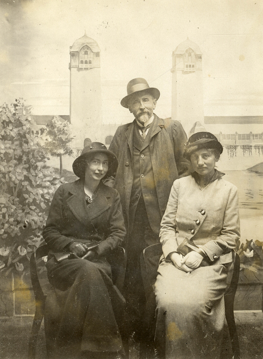 Gruppeportrett. Mann og to kvinner foran fotobakgrunn som forestiller bygning med to tårn. Fra Jubileumsutstillingen 1914.