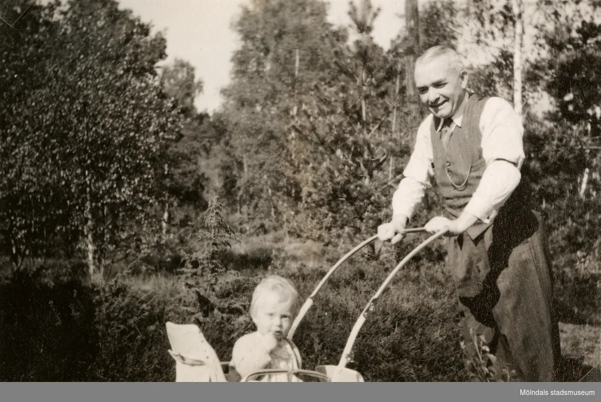 Carl Krantz (1880 - 1956) är på promenad med dotterdottern Eva Pettersson (född 1944, gift Kempe) som sitter i kärran, Stretered år 1945. Carl Krantz var skomakare och undervisade vid Stretereds skolhem.