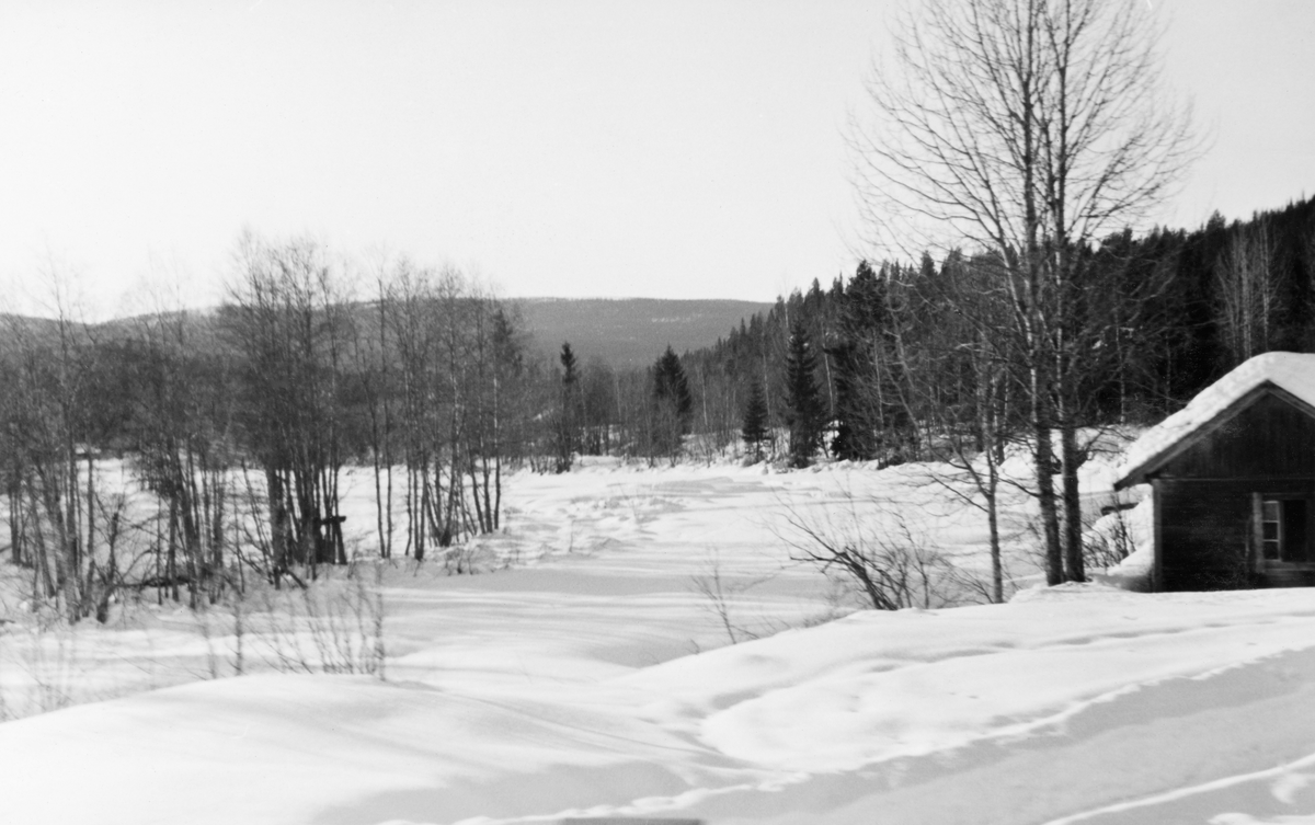 Vinterlandskap ved elva Hovda, ei sideelv som renner inn i Glomma fra vest, og som fungerer som kommunegrense mellom Stor-Elvdal og Åmot i Hedmark. Dette fotografiet ble tatt fra et togvindu i februar 1938. Det viser et snødekt landskap hvor vi så vidt kan ane det islagte elveløpet som en forsenkning i snøflata. Langsmed elvebredden sto det lauvskog, antakelig oretrær, med granbestand bak. I forgrunnen var det ei åpen flate, hvor vi ser en snøplogkant og et hushjørne, antakelig på garden Søre Sætres grunn.