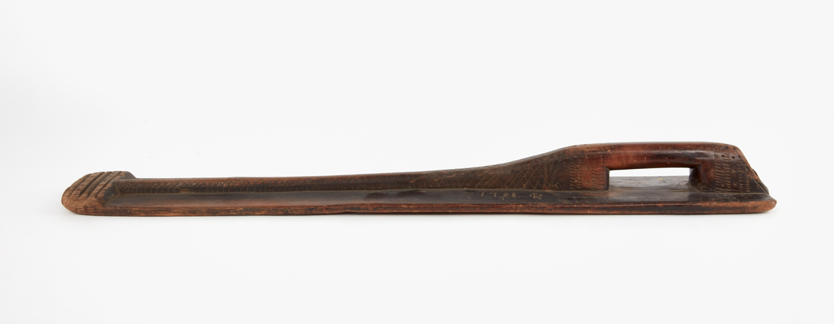 Mangeldon av trä med kavel. Handtag utsparat i ett stycke med brädan. Handtagets förlängning bildar mittås, som har ristad ornering i rutmönster. 
Märkt med initialer och bomärke samt år 1791. Rak typ.