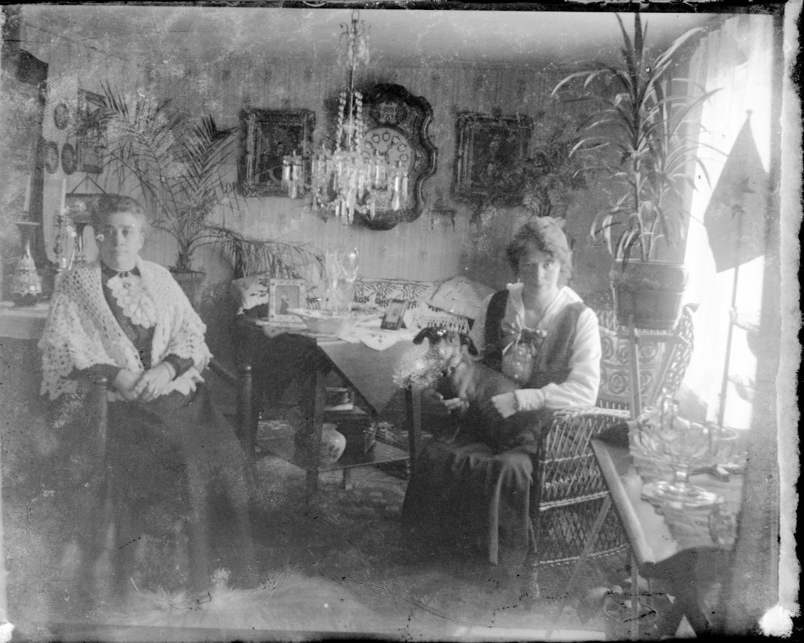 Två kvinnor i en hemmiljö. Lågt i tak med kristallkrona över ett bord. Väggklocka mellan porträtt. En äldre kvinna sitter i en munkstol och en yngre, med tax i knät, i en korgstol.