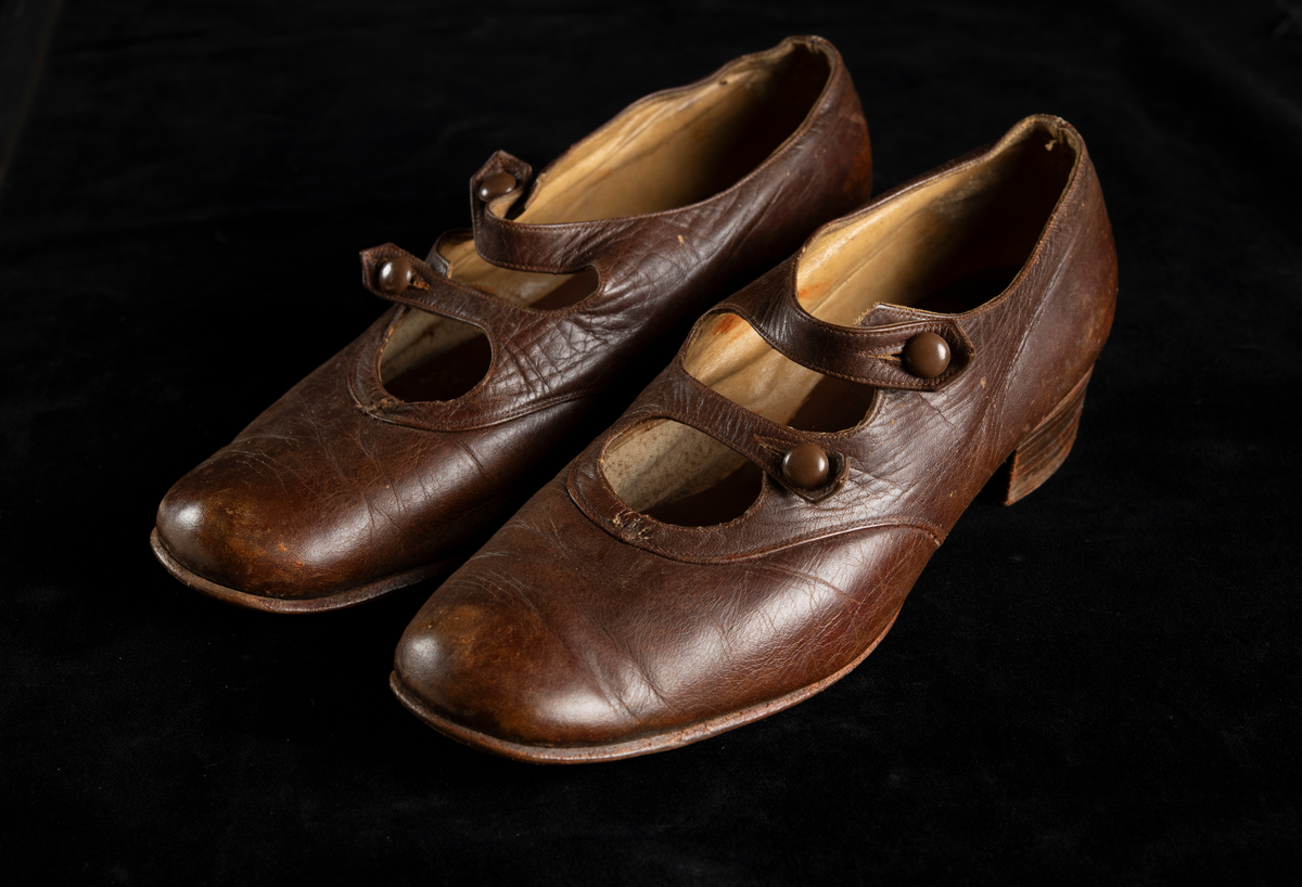 Ett par skor av brunt skinn. Flickmodell i storlek 35. Klackhöjd 25 mm. Dubbla slejfar som knäpps med små bruna plastknappar.