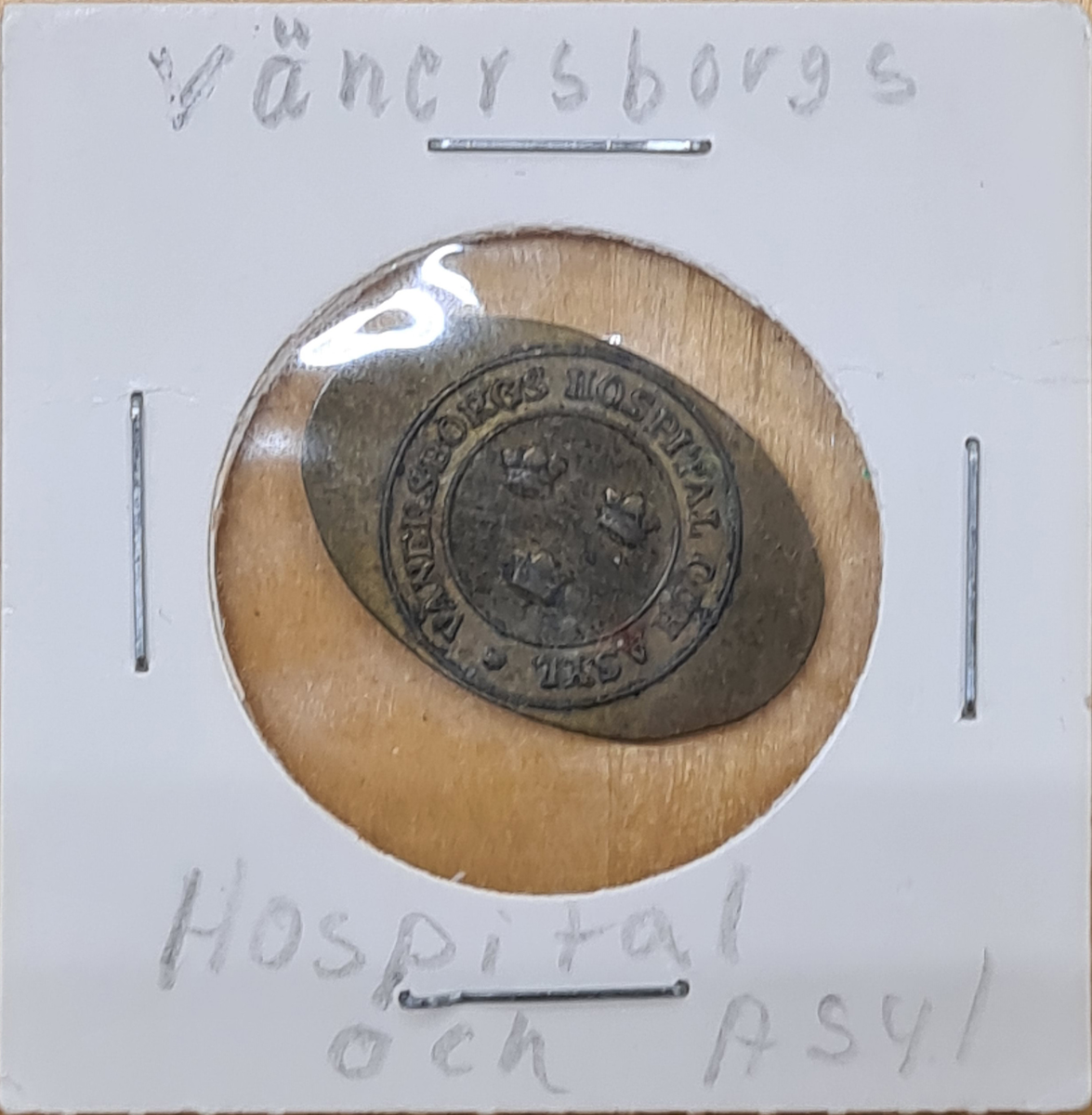 Polett i koppar. Poletten är oval med en stansad ring med tre kronor sär det står: Vänersborgs Hospital och Asyl.