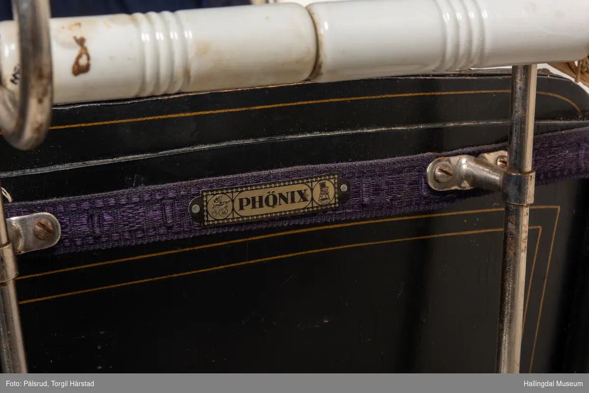 Sort barnevogn fra 1920-tallet. Gullborder på sidene og mørk blå forheng forran "hetta". Av merke Phönix.