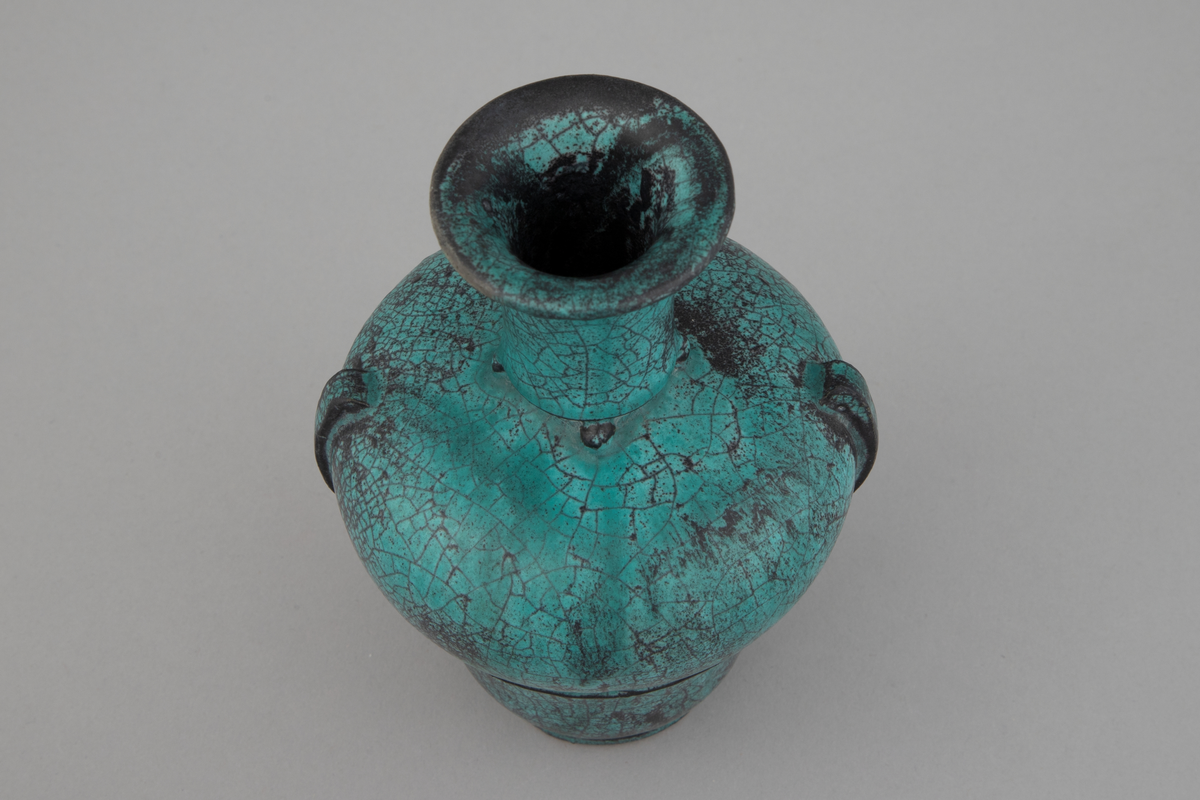 Liten vase med brede skuldre og smal hals. Vasen smalner inn ned mot foten. Den er glasert med grønn askeglasur med svarte partier, halvmatt og krakelert.