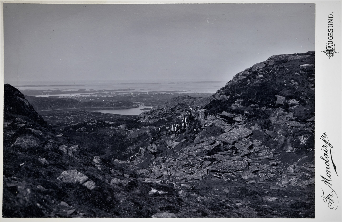En gruppe menn og kvinner på tur på vei opp mot Steinsfjellet. I bakgrunnen ses Eivindsvatnet og Haugesund.