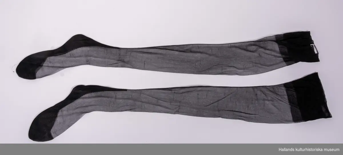 Sorgedräkt. a) Klänning. Svart klänning i syntetmaterial. Klänningen är knälång och har knappar fram. I sidan är det en dragkedja. Längd 97 cm. Bredd 70 cm. Ärmlängd 52 cm. 
b) Sorgflor av ett runt stycke svart tyll bandkantat med svart tyg som sytts fast i zig-zag. Måtten i måttfältet avser sorgfloret. 
c) Skärp, svart till sorgdräkt. Hålls ihop med hjälp av en hake. Tillverkat av syntettextil. Mått: längd 79 cm, bredd: 1,5 cm. 
d-e) Skor i storlek "3H". Märkta: "Cats Gyllene Gripen, Svensk tillverkning". Skorna är gjorda av svart skinn. På häl och tå är det svart lack vilken är dekorerad med hål. Även den halvhöga klacken är klädd med lack. 
Mått: längd 32 cm, bredd 8 cm, höjd 9 cm.
 f) Strumpor. Svarta tunna strumpor som skall hållas uppe med strumpeband. Tillverkade av syntettextil. Längd 100 cm. Bredd 16 cm. 
(Klassifikation enl VMF realnummerkatalog: 6:1.5, 6:1.15, 6:1.16, 39:3)