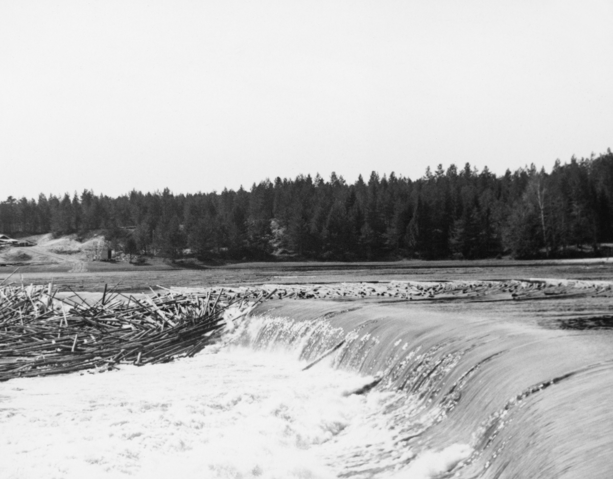 Kraftverksdammen ved Skjefstadfossen i Heradsbygda i Elverum.  Fotografiet er tatt i april 1942, etter at vårflommen og tømmerfløtinga hadde begynt å sette sitt preg på Glommavassdraget.  Bildet er tatt fra elvas østre bredd mot elveleiet like nedenfor dammen, der det hadde lagt seg tømmervaser på steinører som kun var dekt av et grunt vannlag.  I bakgrunnen til høyre ser vi inntaksdammen, der turbinrørene til kraftverket, som vi ser litt mer sentralt i bildet (og litt lengre nede ved elvas vestbredd).  Landskapet i bakgrunnen preges av barskog.  I Glomma fellesfløtingsforenings album nr. 13 er dette fotografiet montert tett inntil SJF. 1989-03951, som en slags vidvinkelmontasje.  Vinteren før dette fotografiet ble tatt foregikk det anleggsarbeid på dammen (jfr. SJF. 1989-03947-49). 