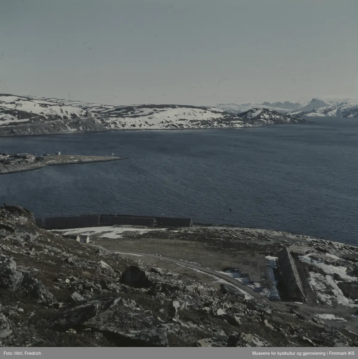 Bildet er fotografert fra Vardfjellet ovenfor Meland. I forgrunnen ser man fiskehjeller der fisken henger til tørk. Utsikten går over Rossmollbukta og innseilingen til Hammerfest havn til veien på Salenfjellet opp til Storsvingen og Rypefjellet i bakgrunn. Det ligger fortsatt snø.