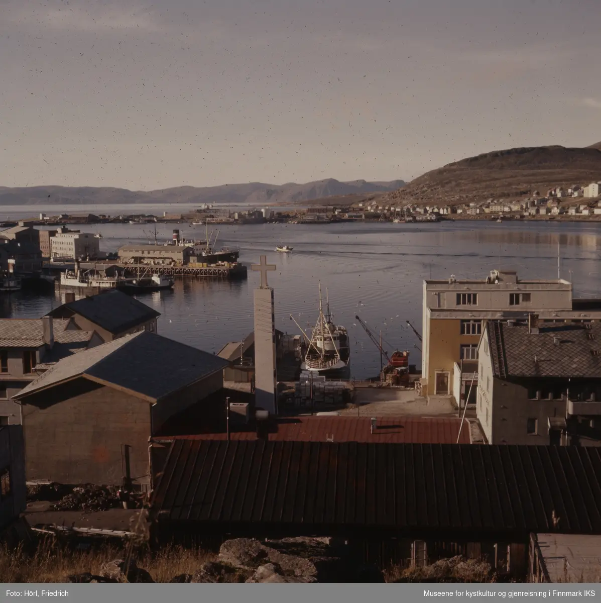 Utsikt fra Salenfjellet med tårnet til Den katolske kike i forgrunn, Hammerfest havn i midten og bydelen Fuglenes i bakgrunn. I havna ligger båter og man kan se dampskipskaia og en del av Findusfabrikken med kaianlegg og kraner.