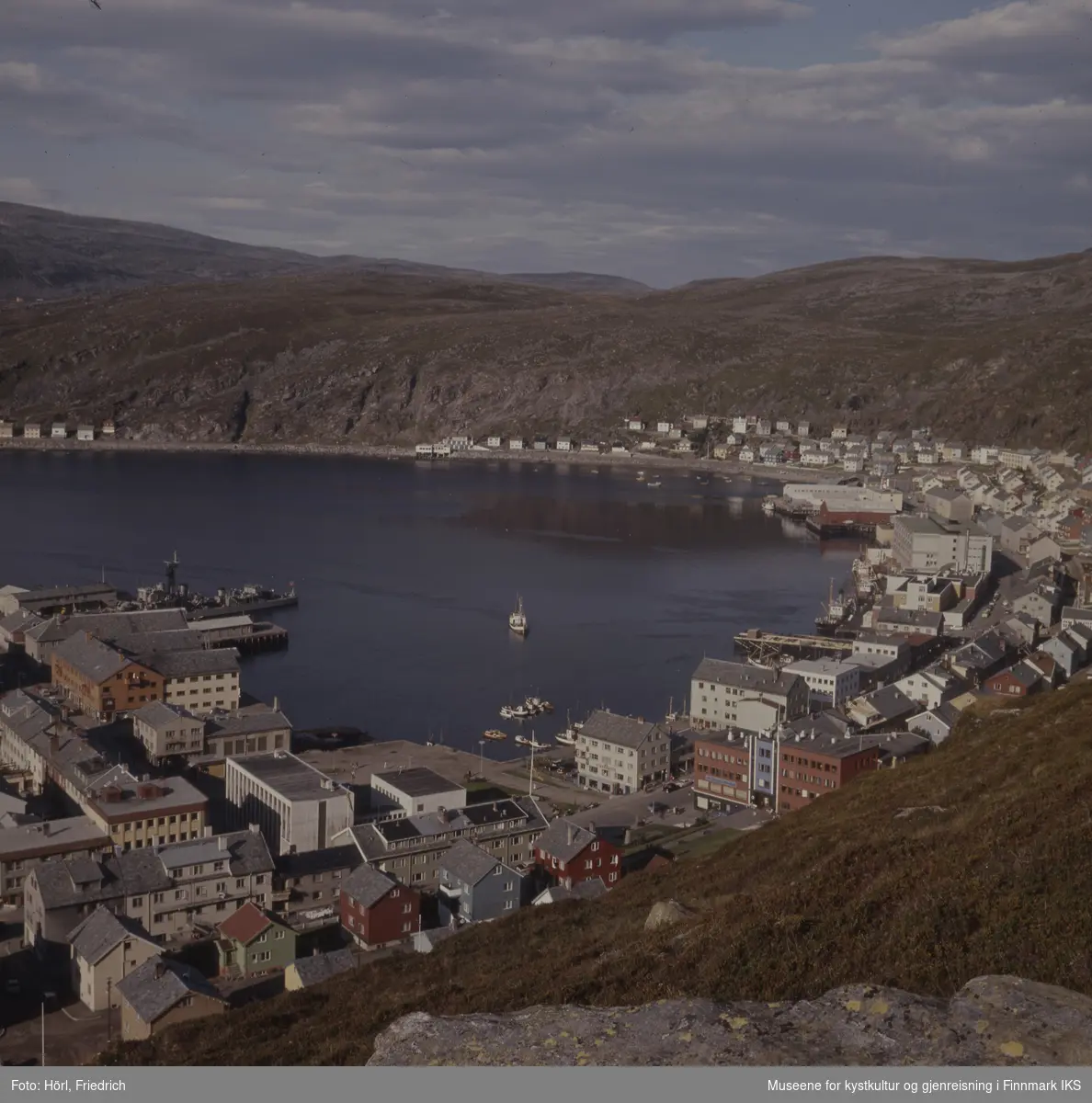 Det er sommer i Hammerfest og fra Salenfjellet ser man utover byens sentrum med rådhus, bankbygg og Findusfabrikken helt bort til Hauans materialandel ved munningen til Storelva og bydelen Molla bak den. Byen er gjenreist og fargerike hus med skifertak preger bybildet. I havna liger det ulike typer båter. Marinefartøyet på bildet er en norsk fregatt, mest sannsynlig KNM Tromsø eller KNM Haugesund.