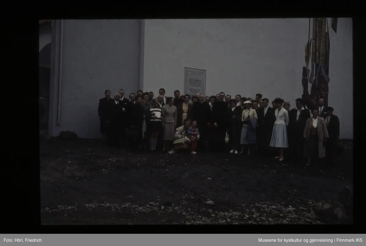 En gruppe personer i ulik alder står foran Den katolske kirke St. Mikael i Hammerfest i 1958. De er festkledde. Bak dem ser man deler av Karl Manningers veggmosaikk.