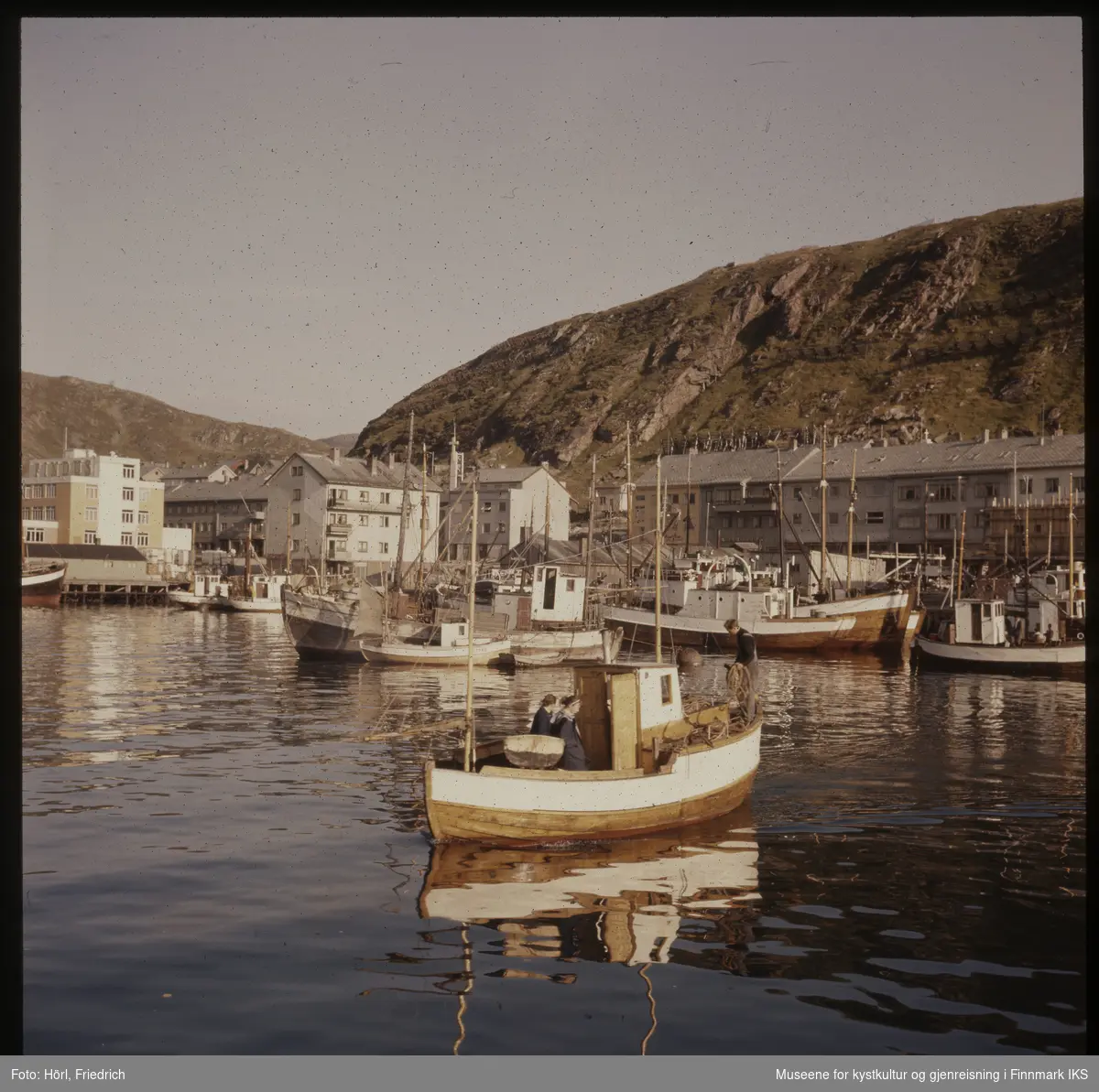 I Hammerfest havn ligger mange fiskeskøyter. I forgrunnen ser man en kvinne og to  menn på en fiskebåt. I bakgrunnen ser man Salenfjellet og gjenreisningsbebyggelse foran i Hammerfest sentrum. Helt til venstre i bilde ser man dampskipskaia og en del av Findusfabrikken. Bildet er tatt i 1957 eller 1958.