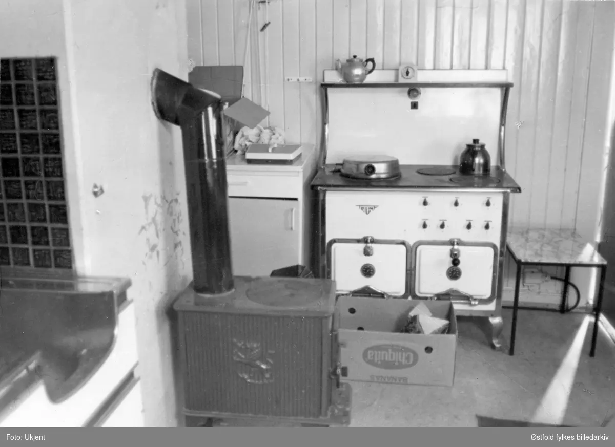 Gården Alvim nordre i Tune, fotografert februar 1977.
Fra våningshus,kjøkkeninteriør med magasinkomfyr av merket Triumf, vedovn, badevekt, kjøkkenskap, bord, ved i pappeske, kaffikanne.
Våningshus bygd omrking 1780, restaurert 1955.